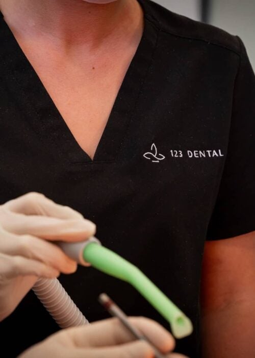 Gloved clinician in black 123 Dental scrubs holding a suction tube and a hand instrument
