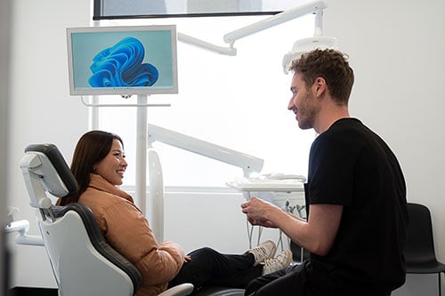 Dentist in black seated on a stool chatting with a smiling patient in the dental chair