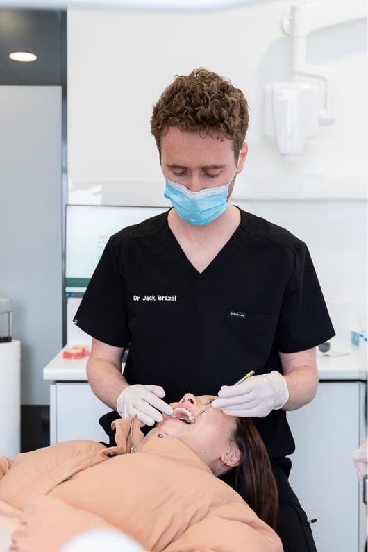 Masked dentist examining a reclined patient's teeth with a mirror and probe in the surgery