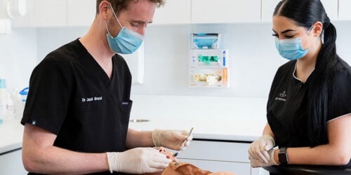 Masked dentist and assistant in black scrubs examining a reclined patient's teeth with a mirror