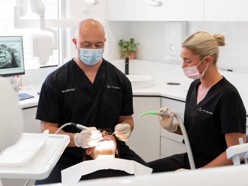 Masked dentist and assistant in black scrubs treating a patient reclined in the chair