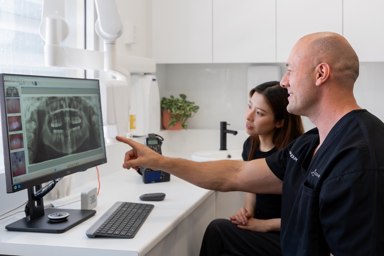 Dentist pointing to a panoramic dental X-ray on screen while a patient looks on