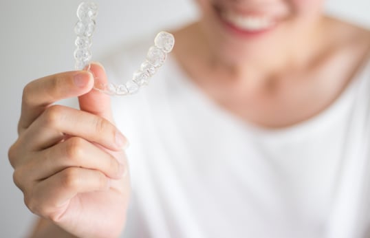 Smiling person in a white t-shirt holding up a clear removable aligner between finger and thumb
