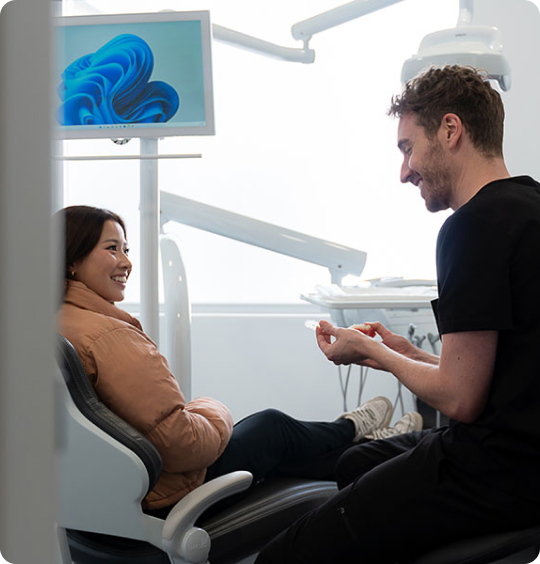 Patient smiling up from the dental chair as a dentist in black talks with her