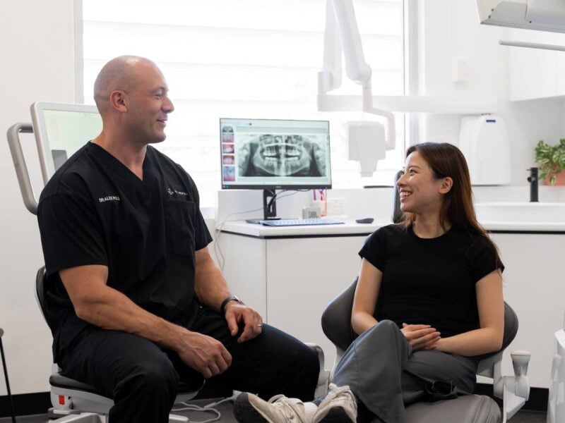 Dentist chatting with a smiling patient in the chair, a panoramic x-ray on the screen behind